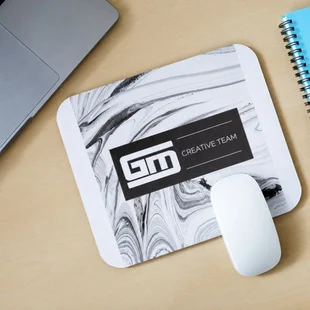 White mouse on a marbled mouse pad labeled "Creative Team" next to a laptop and a spiral notebook on a wooden desk.