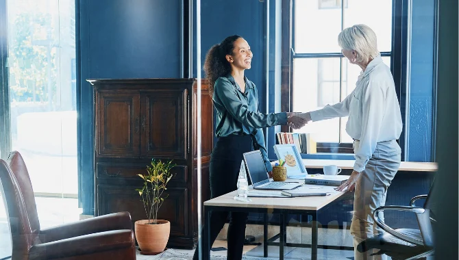 Two people shaking hands in a blue-walled office, with a wooden bookcase and a desk holding a laptop and folders.