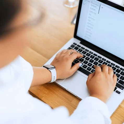 Person typing on a laptop with a word document open, wearing a smartwatch, sitting at a wooden desk.