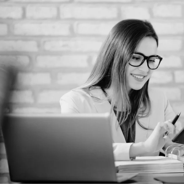 Smiling person with glasses uses a tablet at a desk with a laptop. Brick wall background.