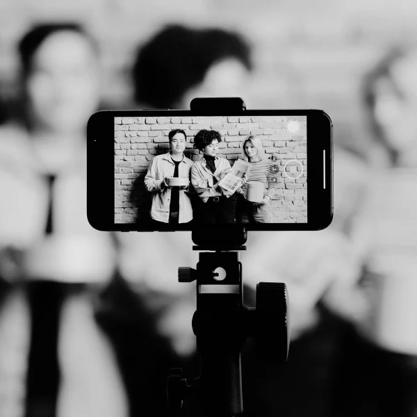 Smartphone on tripod capturing three people holding beverages, standing against brick wall. Black and white photo, background blurred.