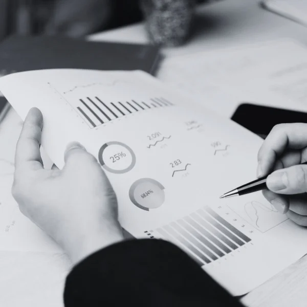 Hands holding a paper with graphs and charts; one hand points with a pen. Black and white theme.