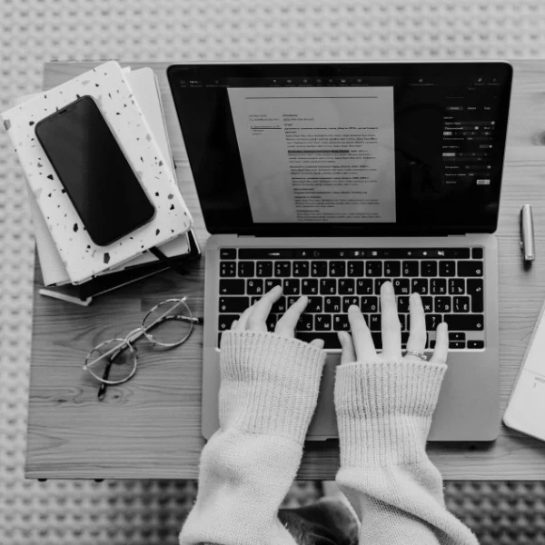 Hands typing on a laptop, a resume draft visible. Glasses, phone, and notebooks beside, sweater sleeves pulled over hands.