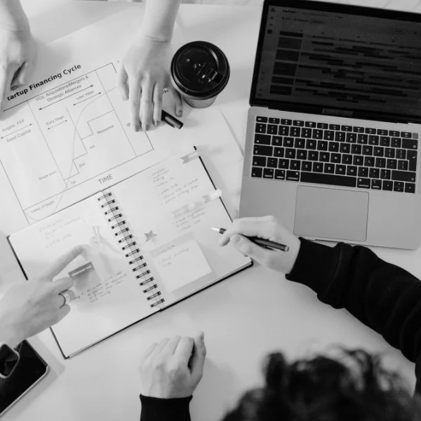 Team collaborating over graphs and notes, with open laptop and coffee cup on desk. Hands point and write in a notebook.